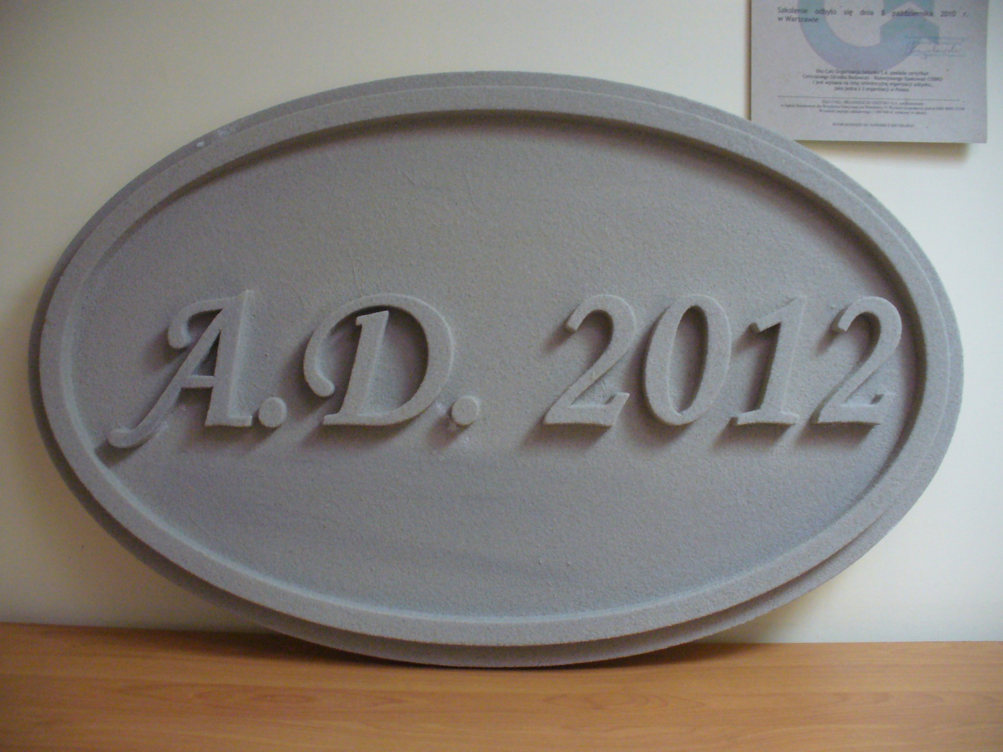 Szara, owalna tablica z napisem 'A.D. 2012' wykonanym w reliefie, umieszczona na drewnianym blacie, z widocznym fragmentem certyfikatu na ścianie w tle.
