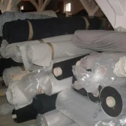 Stos rolek tkanin w magazynie, widoczne tkaniny poliestrowe w różnych kolorach, niektóre zapakowane w folię, na tle drewnianych belek stropowych.