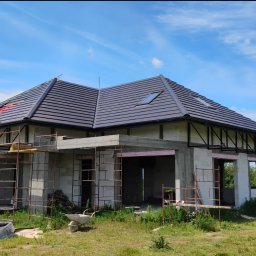 M-BUD Usługi Remontowo-Budowlane - Dom w stanie surowym otwartym z ciemną dachówką, widoczne rusztowania i prace na dachu. Błękitne niebo z chmurami w tle. Etap budowy z użyciem białych bloczków.