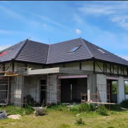 Dom w stanie surowym otwartym z ciemną dachówką, widoczne rusztowania i prace na dachu. Błękitne niebo z chmurami w tle. Etap budowy z użyciem białych bloczków.