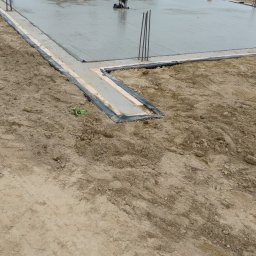 M-BUD Usługi Remontowo-Budowlane - Wylany fundament pod budynek z widocznym zbrojeniem i izolacją, w tle żółty budynek gospodarczy i drzewa. Prace fundamentowe w toku, widoczni pracownicy.