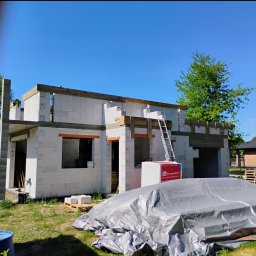 M-BUD Usługi Remontowo-Budowlane - Budowa domu z betonu komórkowego: ściany w trakcie wznoszenia, widoczne nadproża i rusztowanie. Część materiałów budowlanych zabezpieczona folią. Słoneczny dzień.