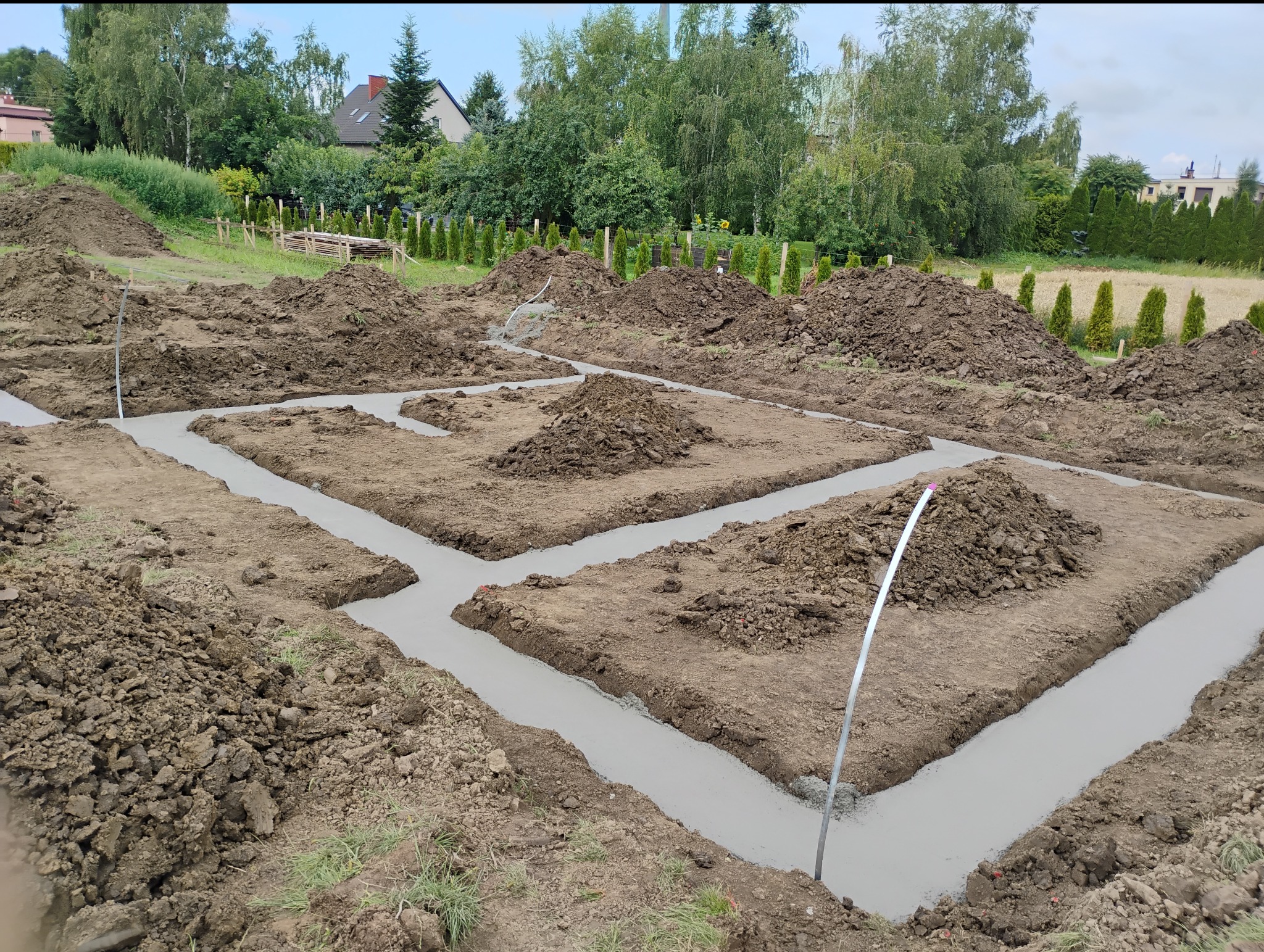 Zalane fundamenty pod budowę domu jednorodzinnego. Widoczne wykopane rowy i wylany beton. Na ziemi leżą pryzmy ziemi. W tle drzewa i budynek.