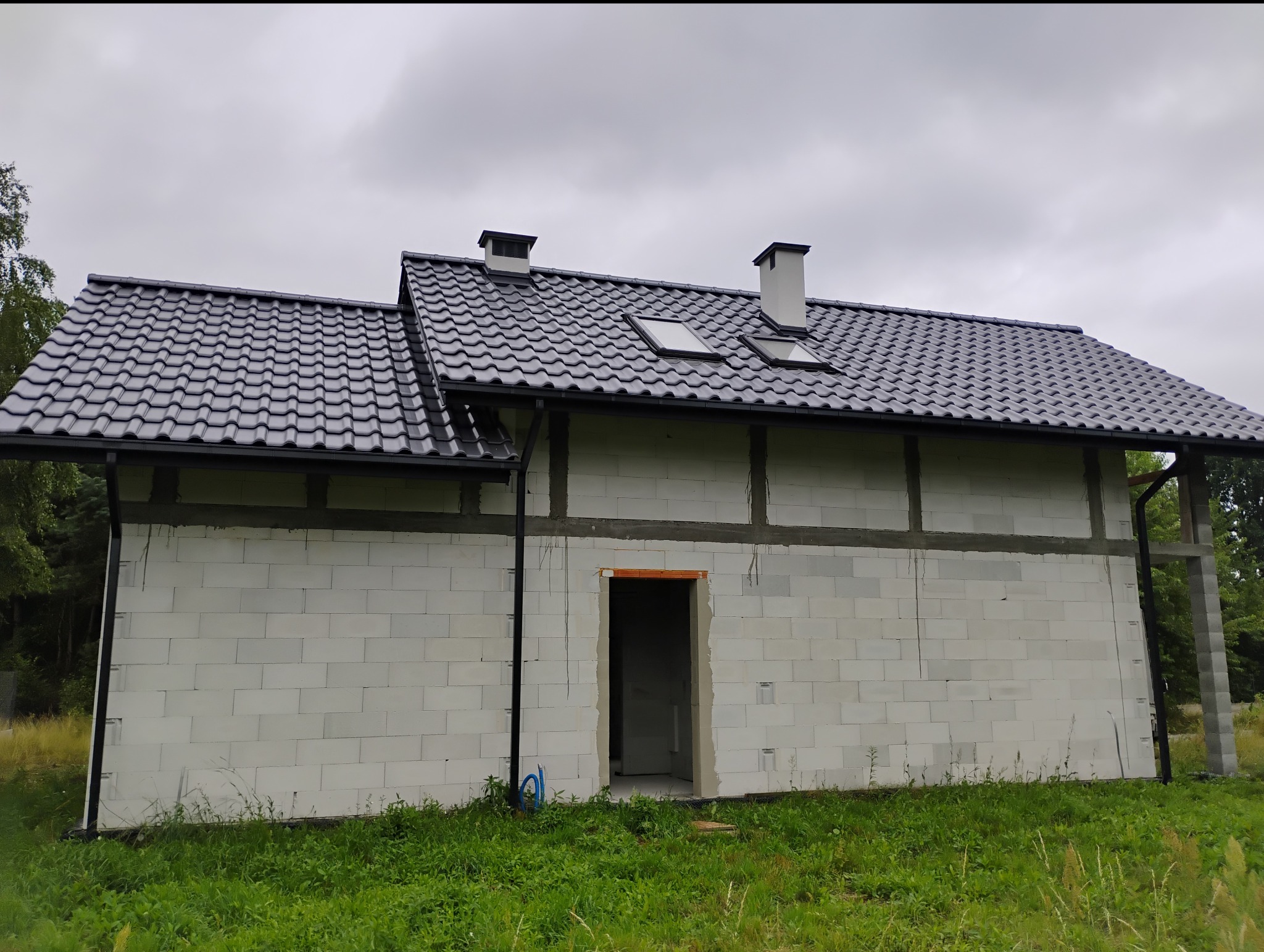 Surowy dom z białego pustaka z ciemną dachówką, widoczne kominy i okna dachowe. Budynek w trakcie budowy, otoczony zielenią.