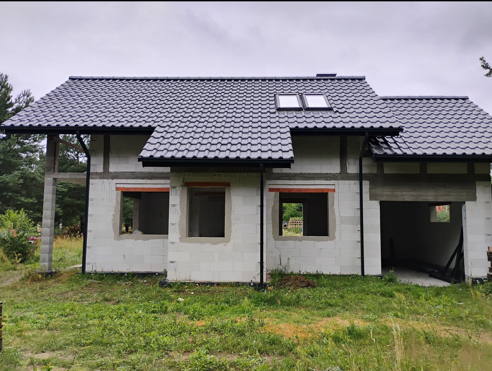 Niedokończony dom z białego betonu komórkowego z ciemnoszarą dachówką i oknami dachowymi, otwarty garaż, widok na zieleń przez okna.
