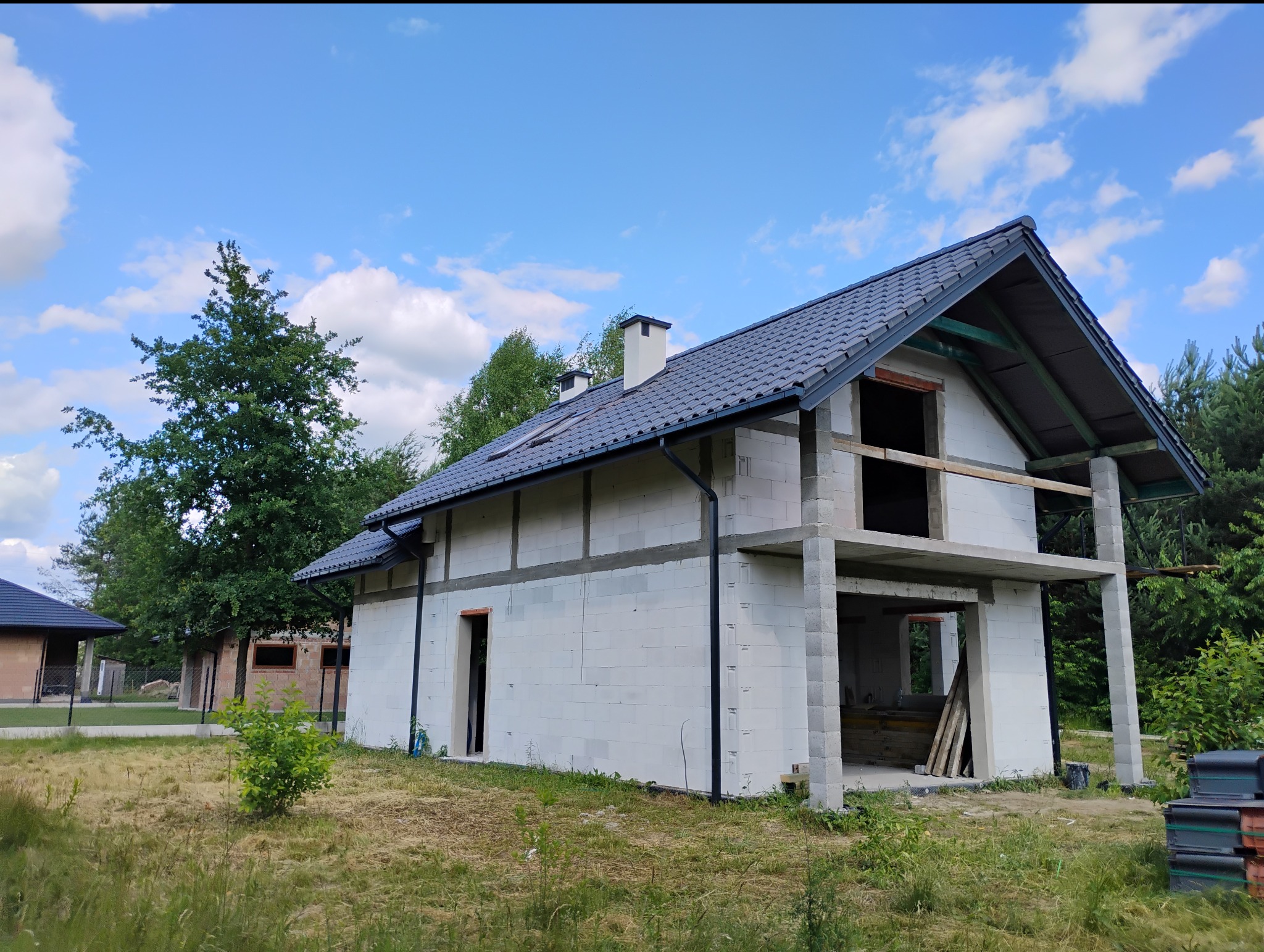 Niedokończony dom z białych bloczków, z szarym dachem i widoczną więźbą. Częściowo otynkowany, z otworami okiennymi i drzwiowymi. W tle zieleń i błękitne niebo.