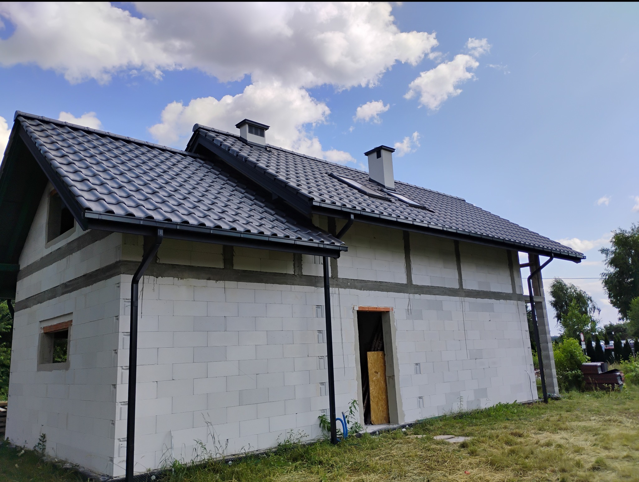 Niedokończony dom z białych bloczków, czarny dach z dachówki ceramicznej, widoczne kominy i rynny. Błękitne niebo z chmurami w tle. Etap budowy.