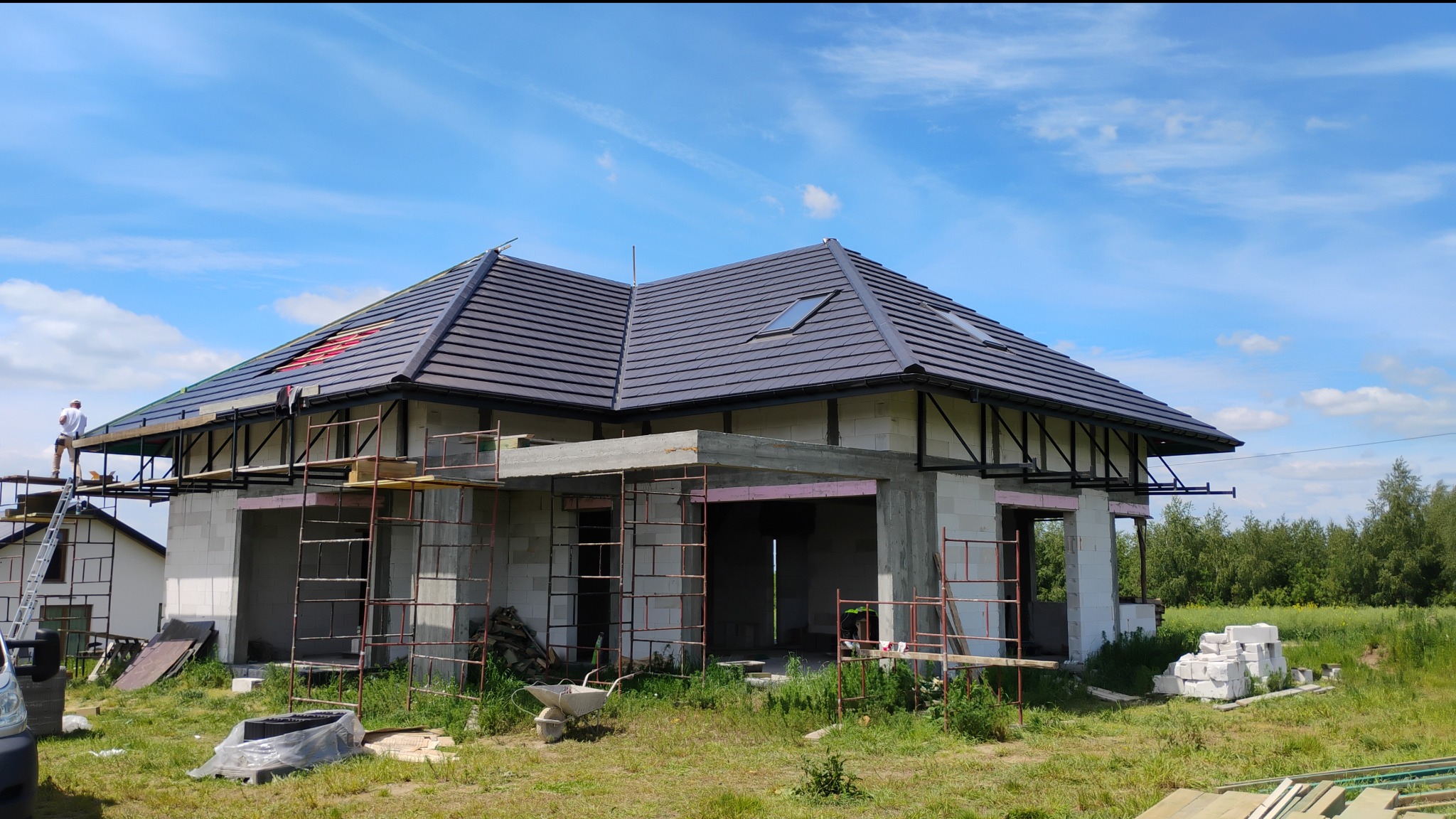Dom w stanie surowym otwartym z ciemną dachówką, widoczne rusztowania i prace na dachu. Błękitne niebo z chmurami w tle. Etap budowy z użyciem białych bloczków.