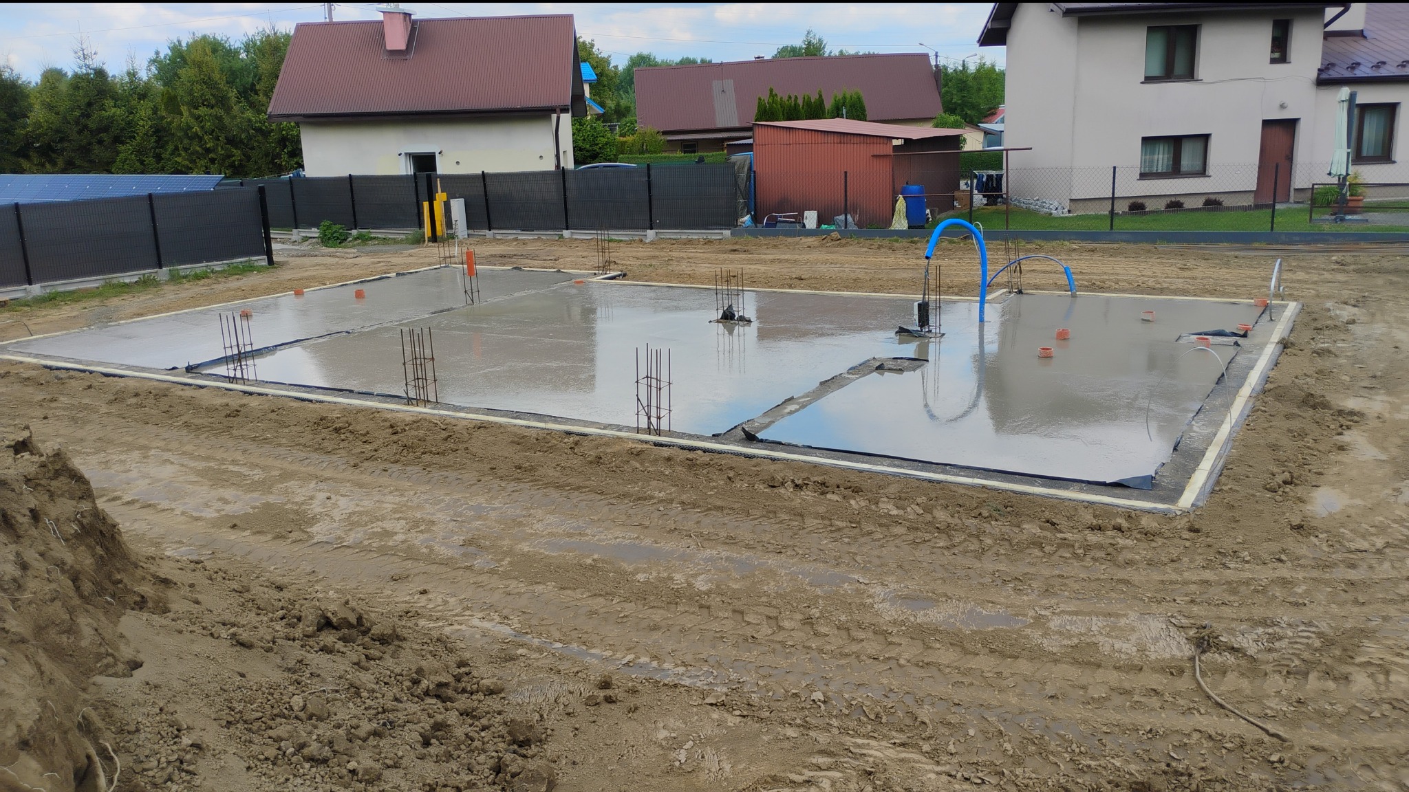 Świeżo wylany fundament z wystającymi prętami zbrojeniowymi, otoczony czarną folią izolacyjną. W tle zabudowa jednorodzinna.