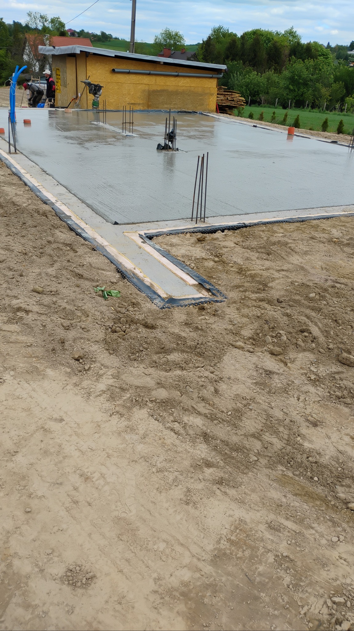 Wylany fundament pod budynek z widocznym zbrojeniem i izolacją, w tle żółty budynek gospodarczy i drzewa. Prace fundamentowe w toku, widoczni pracownicy.