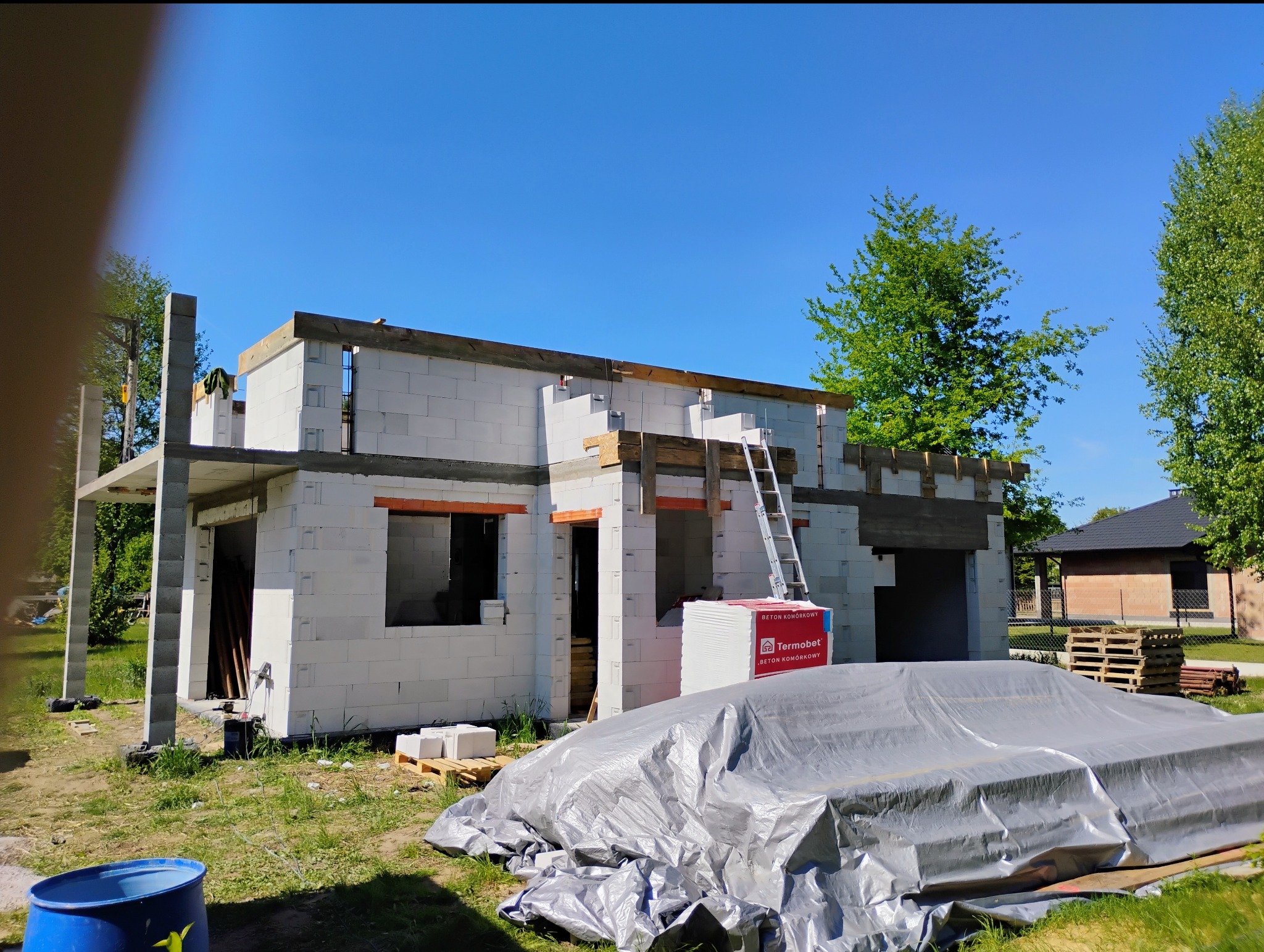 Budowa domu z betonu komórkowego: ściany w trakcie wznoszenia, widoczne nadproża i rusztowanie. Część materiałów budowlanych zabezpieczona folią. Słoneczny dzień.