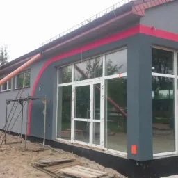 Szary budynek z dużymi oknami, czerwonymi akcentami i rusztowaniem w trakcie prac wykończeniowych elewacji.