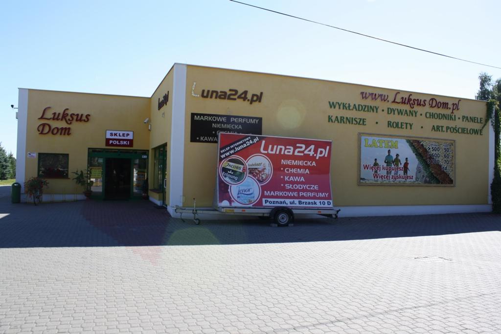 Budynek sklepu z artykułami wyposażenia wnętrz i perfumerii, z szyldami 'Luksus Dom' i 'Luna24.pl', oraz przyczepka reklamowa z ofertą sklepu internetowego Luna24.pl.