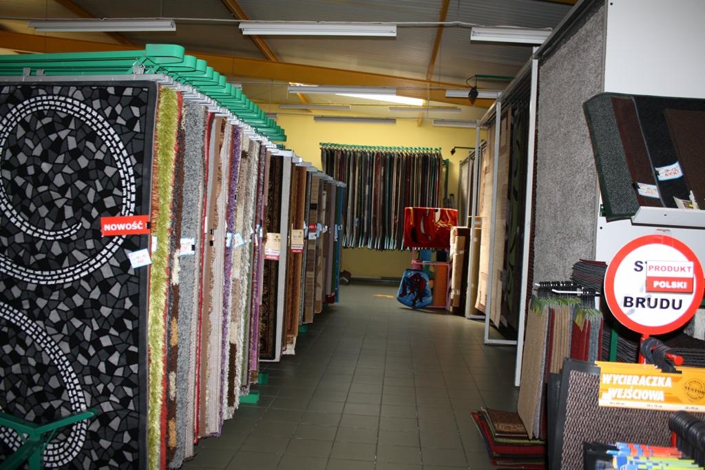 Wnętrze sklepu z dywanami, rzędy dywanów wiszących na metalowych stelażach, widoczne etykiety 'Nowość' na niektórych modelach, w tle ekspozycja zasłon i wycieraczek.