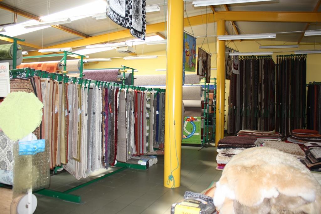 Wnętrze sklepu z dywanami, prezentacja różnorodnych wzorów i faktur dywanów wiszących na ekspozytorach oraz ułożonych w stosy, oświetlenie jarzeniowe, żółte słupy podtrzymujące konstrukcję dachu.