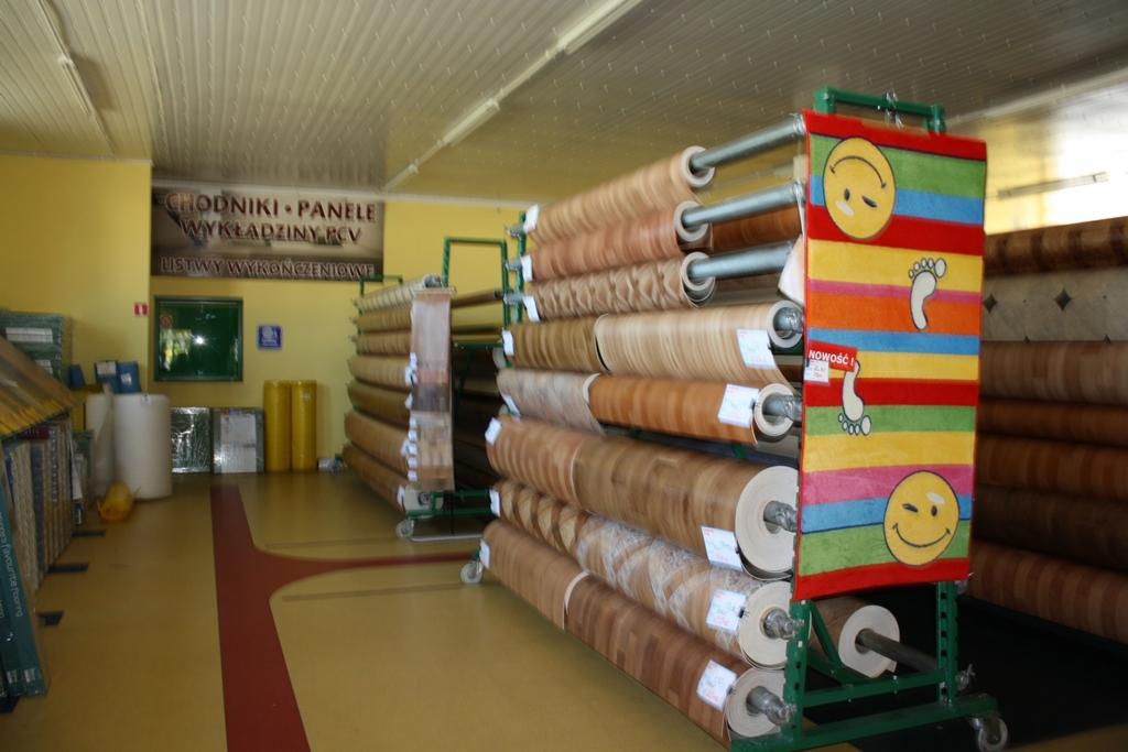 Wnętrze sklepu z wykładzinami podłogowymi w rolkach, widoczne regały z różnymi wzorami i kolorami wykładzin, oraz baner reklamowy z uśmiechniętymi buźkami i stopami.