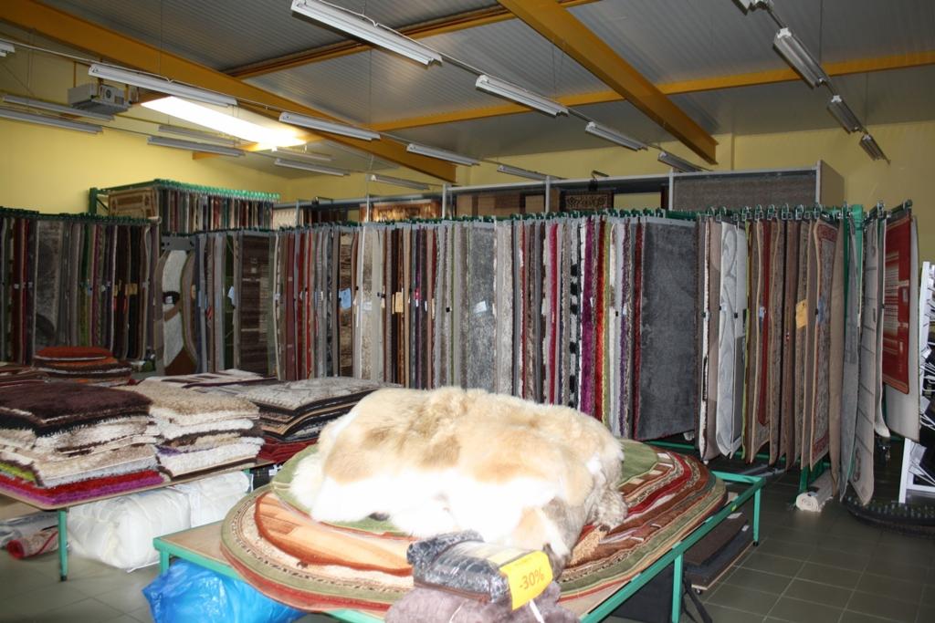 Wnętrze sklepu z dywanami, rzędy dywanów wiszących na ekspozytorach, w pierwszym planie stosy dywanów na stołach, w tym okrągły dywan z długim włosiem, widoczna kartka z informacją o promocji -30%.