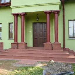 Wejście do domu z kolumnami i schodami wyłożonymi czerwonym piaskowcem, widoczny fragment trawnika z kopcem ziemi.