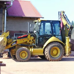 Żółta koparko-ładowarka CAT 432E na placu budowy, z widocznym budynkiem w tle z czerwonym dachem i stertą ziemi.