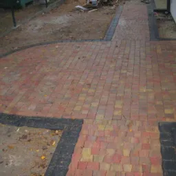 Ułożona kostka brukowa w odcieniach czerwieni i żółci z czarnym obramowaniem, tworząca ścieżkę i okrągły plac przed domem w Łodzi. Widoczne liście na powierzchni.