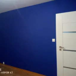 Świeżo pomalowana ściana w odcieniu intensywnego granatu, sąsiadująca z białą, nowoczesną ościeżnicą i drzwiami z poziomymi, szarymi akcentami. Widoczny fragment drewnianej podłogi i biały włącznik...