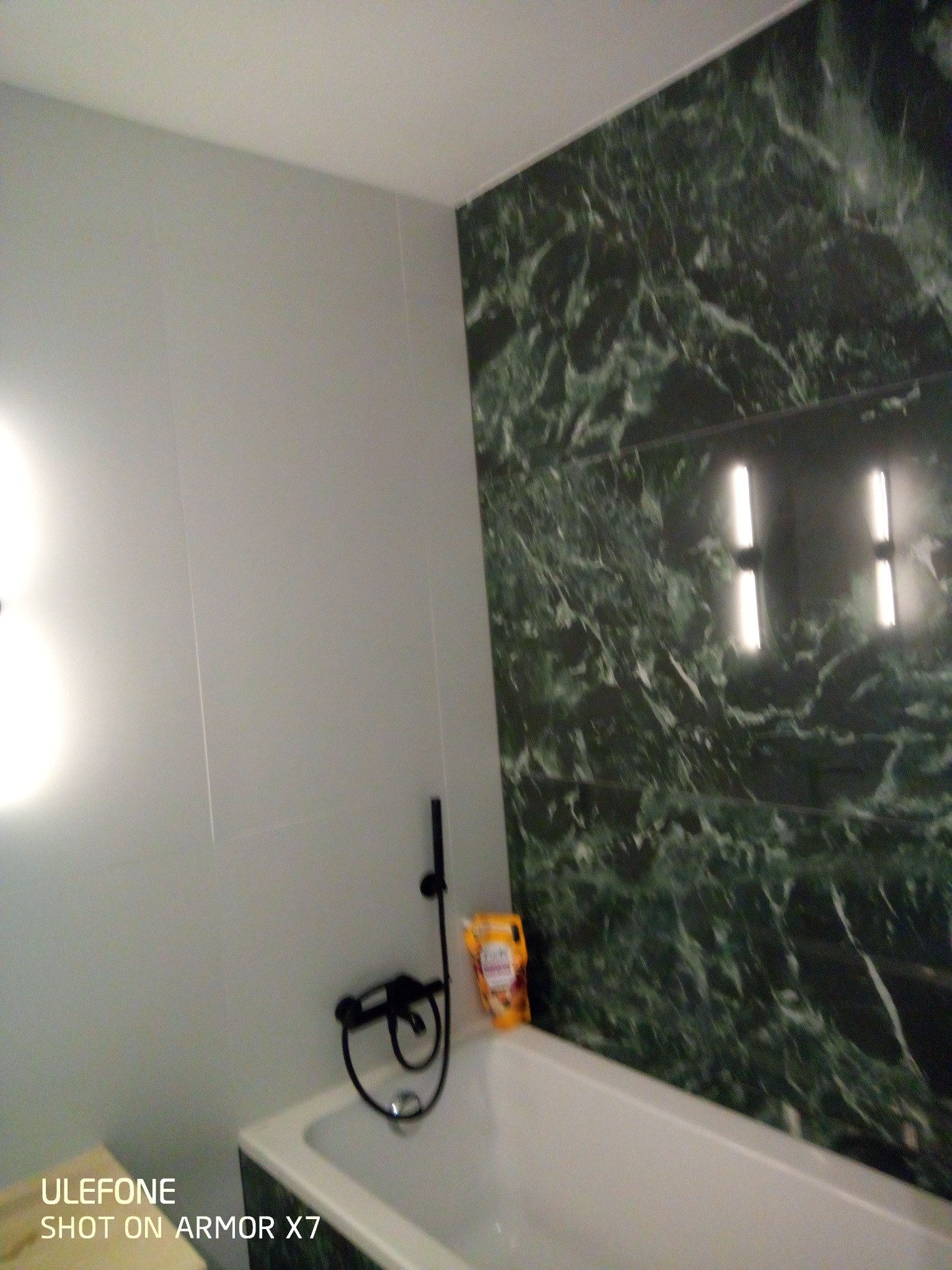 Wyremontowana łazienka z białą wanną, czarną armaturą i ścianą wyłożoną płytkami imitującymi zielony marmur. Na wannie stoi opakowanie kosmetyków.