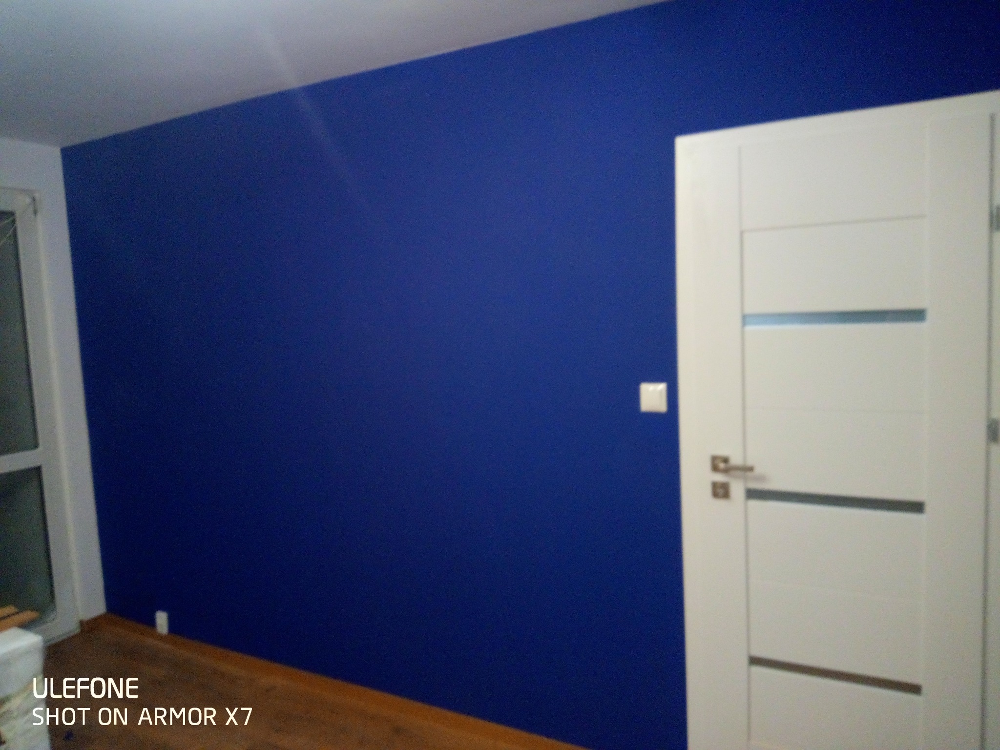 Świeżo pomalowana ściana w odcieniu intensywnego granatu, sąsiadująca z białą, nowoczesną ościeżnicą i drzwiami z poziomymi, szarymi akcentami. Widoczny fragment drewnianej podłogi i biały włącznik...