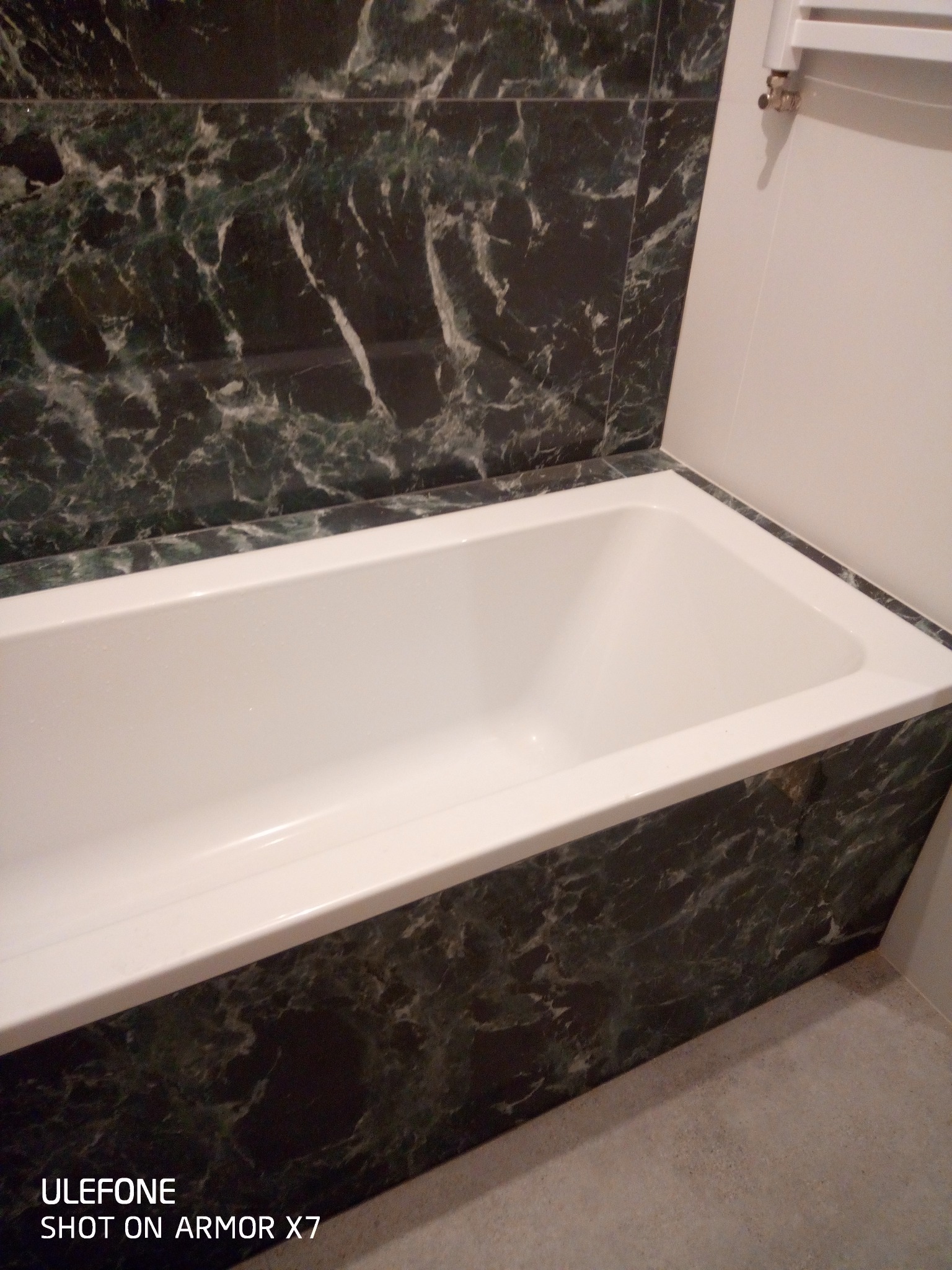 Biała wanna obudowana ciemnozielonymi płytkami imitującymi marmur, z grzejnikiem drabinkowym w tle, po remoncie.