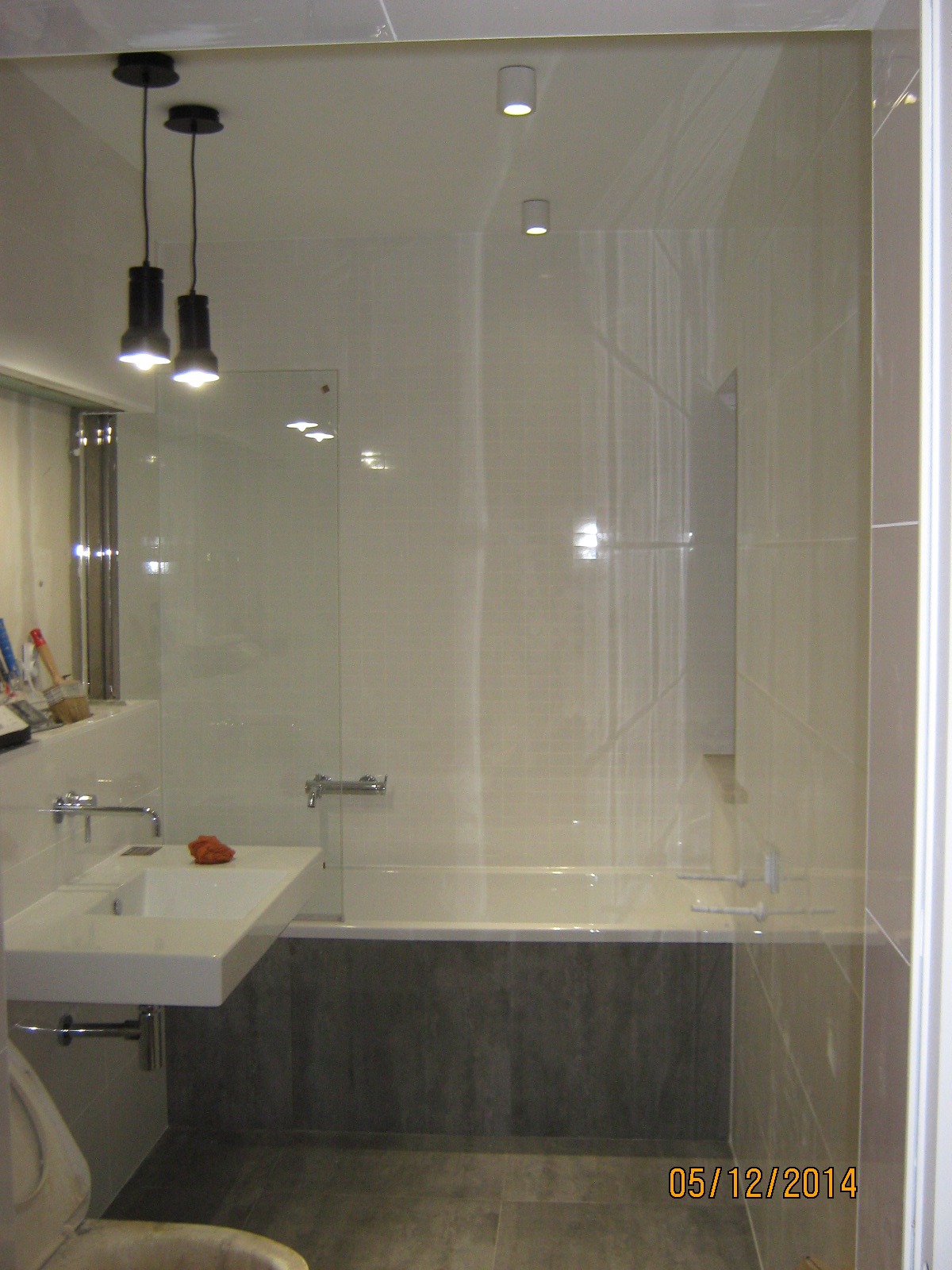 Wykończona łazienka z wanną obudowaną panelami imitującymi beton, umywalką na blacie i szklaną kabiną prysznicową, oświetlona wiszącymi lampami i punktowymi halogenami.