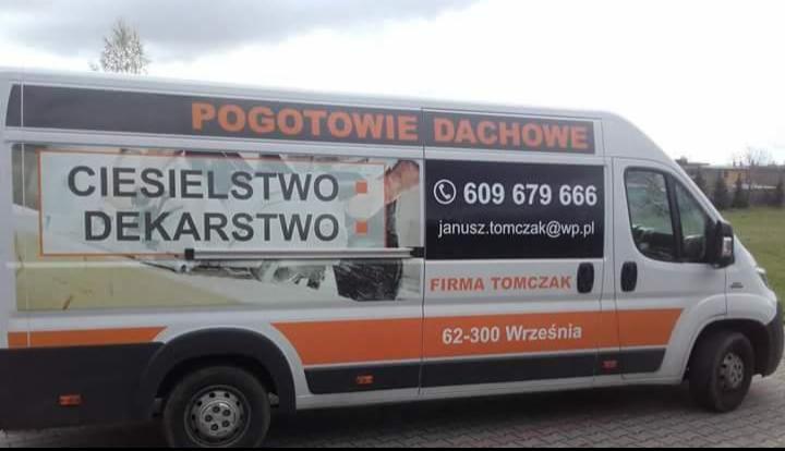 Biały samochód dostawczy z pomarańczowymi akcentami, oklejony reklamą usług ciesielskich i dekarskich, z numerem telefonu i adresem e-mail firmy Tomczak z Wrześni, zaparkowany na kostce brukowej.