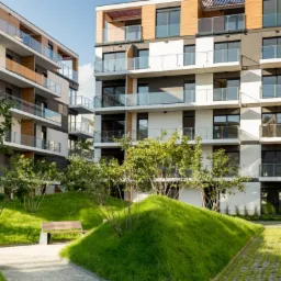 Nowoczesny kompleks apartamentowy z elewacją w odcieniach szarości, bieli i drewna, balkony z przeszklonymi balustradami, otoczony zielenią i ścieżkami spacerowymi.