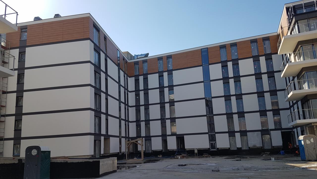 Nowoczesny budynek wielorodzinny z biało-brązową elewacją, widoczne okna zabezpieczone folią, teren budowy w trakcie prac wykończeniowych, przenośna toaleta w rogu kadru.