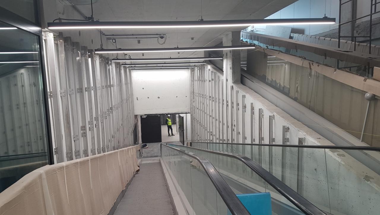 Wnętrze w trakcie prac wykończeniowych z widocznym pracownikiem w kamizelce odblaskowej, fragmentem schodów ruchomych i surowymi ścianami z elementami montażowymi.