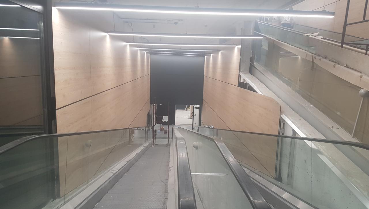 Wnętrze budynku w trakcie wykańczania: widok z góry na schody ruchome z przeszklonymi balustradami, ściany wyłożone panelami drewnopodobnymi i sufit z liniowym oświetleniem LED.