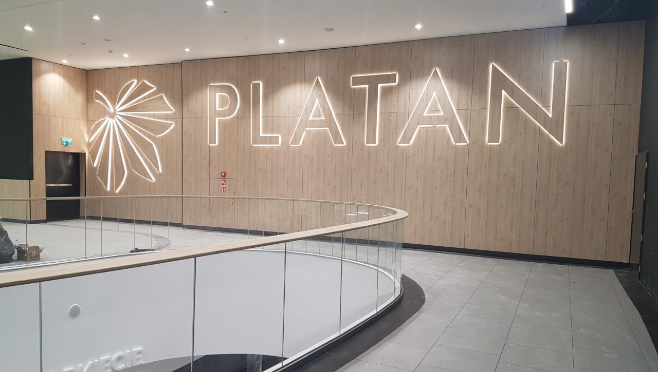 Ściana wewnątrz budynku z drewnopodobnym wykończeniem, na której umieszczono podświetlany napis 'PLATAN' oraz stylizowany liść, widok z antresoli z przeszkloną balustradą.