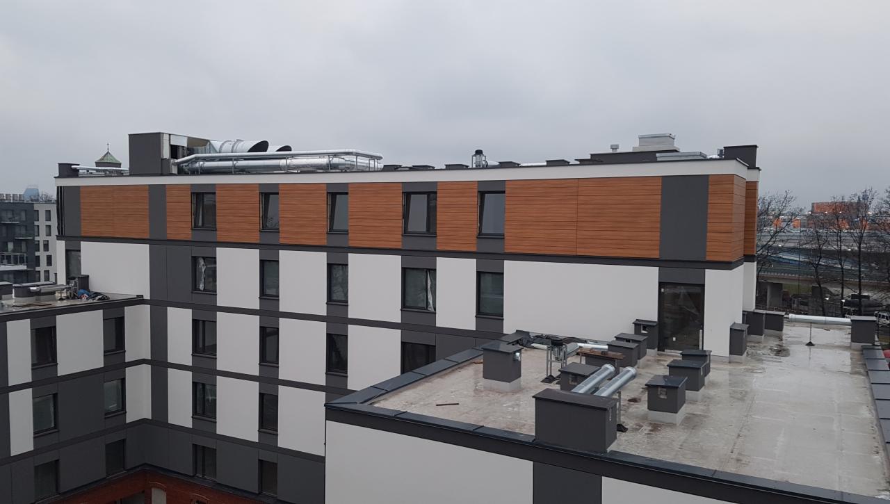 Widok z góry na nowo wykończony budynek wielorodzinny z elewacją w odcieniach szarości, bieli i imitacji drewna, z widocznymi elementami wentylacji na dachu i panoramą miasta w tle.