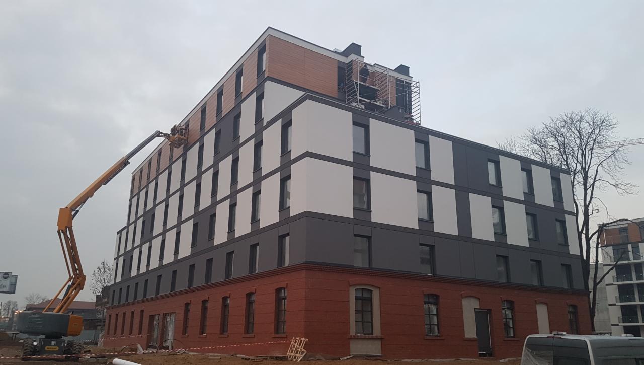 Montaż elewacji budynku wielorodzinnego z wykorzystaniem podnośnika koszowego, widoczna ceglana podstawa i panele elewacyjne w odcieniach szarości, bieli i brązu.