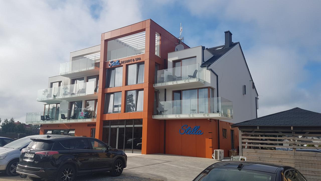 Nowoczesny budynek Stella Resort & Spa w Chłapowie z elewacją wentylowaną w kolorze pomarańczowym i białym, z balkonami z szklanymi balustradami, widoczny z parkingu z samochodami.