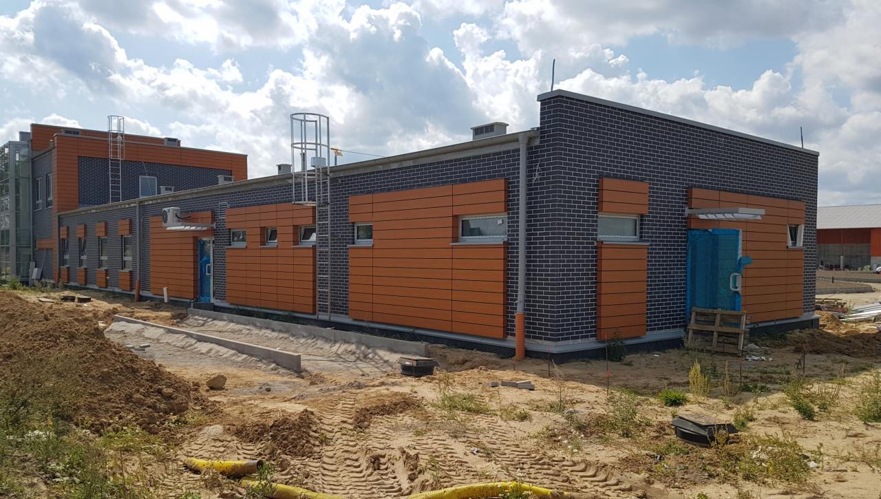 Nowoczesny budynek z elewacją z ciemnej cegły i paneli imitujących drewno, teren wokół w trakcie prac ziemnych.