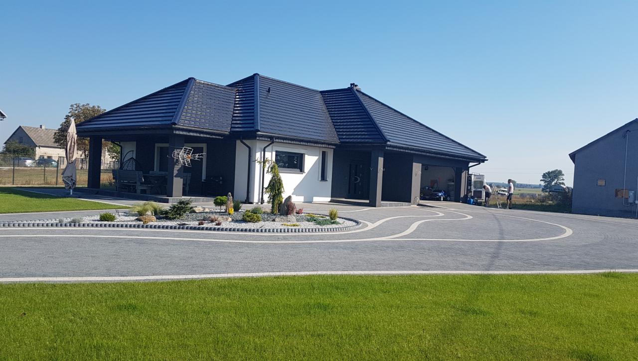 Nowoczesny dom jednorodzinny z ciemną dachówką i grafitową elewacją, biały fragment ściany, ozdobny klomb z iglakami i kamieniami, duży podjazd z szarej kostki brukowej z białymi intarsjami.