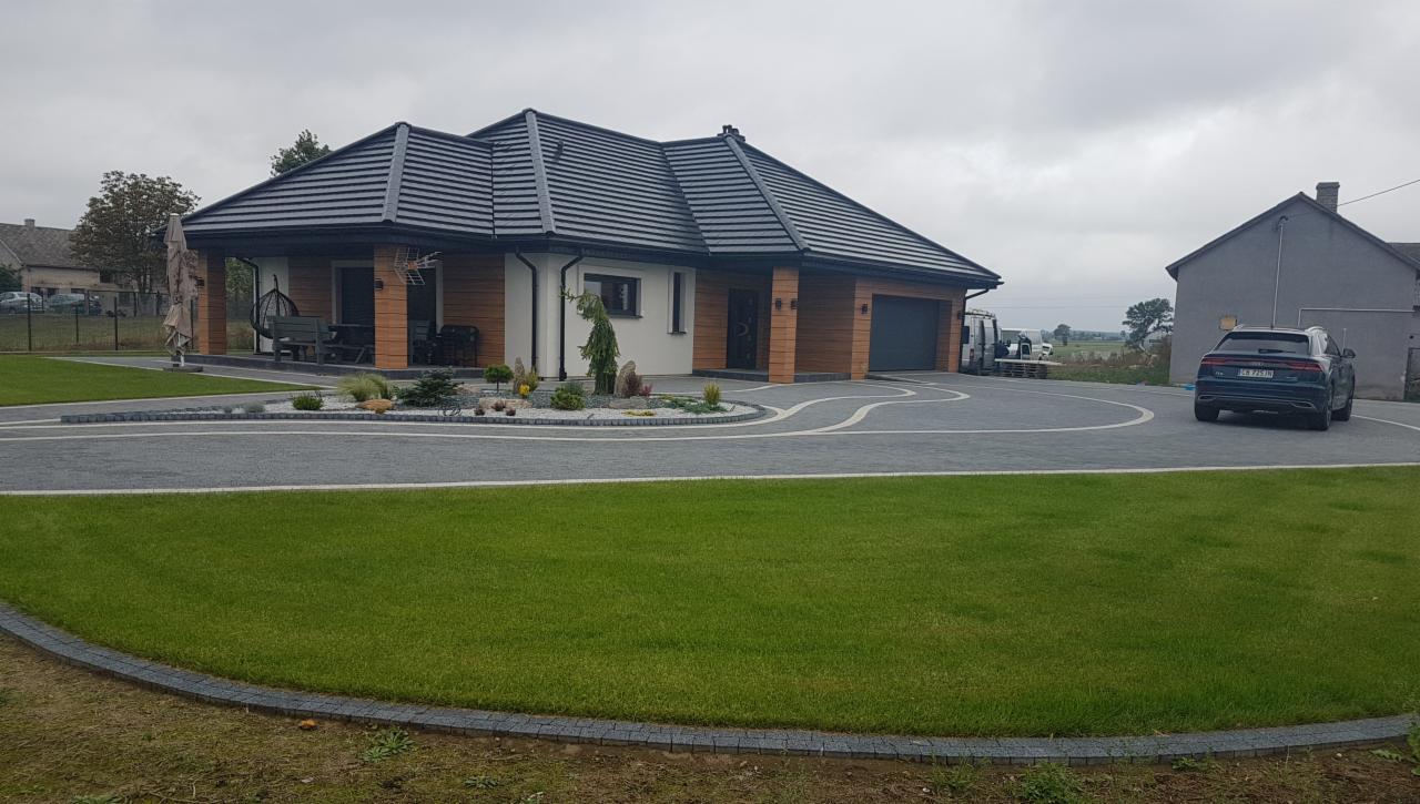 Nowoczesny dom jednorodzinny z ciemną dachówką, drewnianą elewacją i zagospodarowanym terenem wokół, w tle zaparkowany samochód.