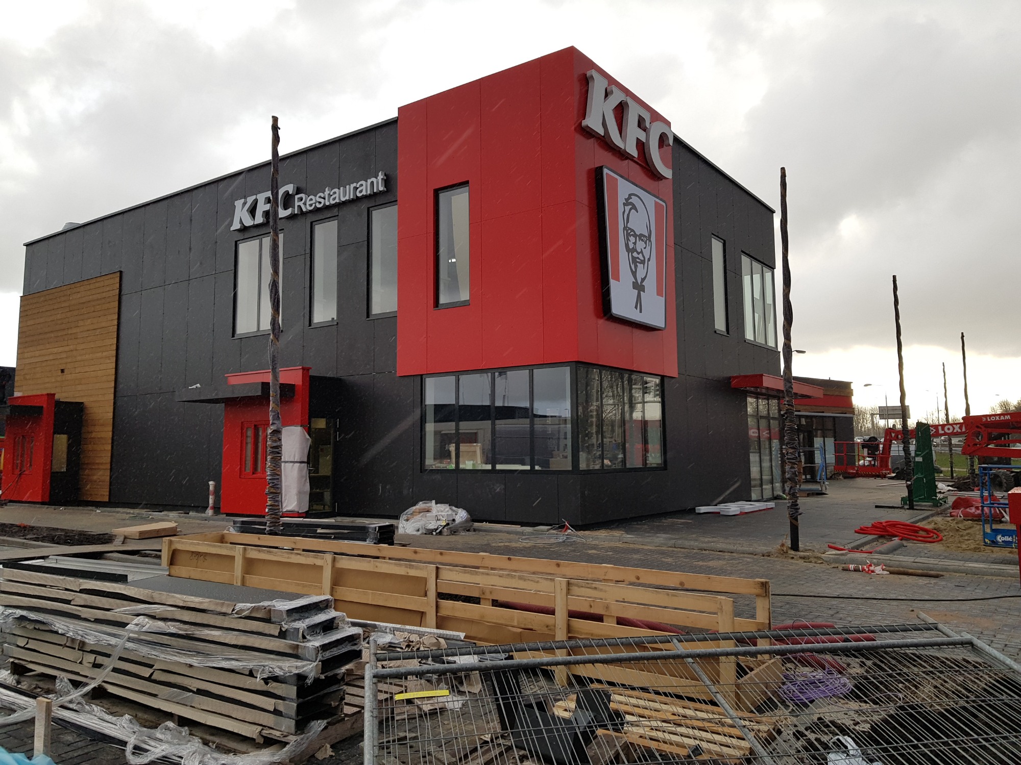 Nowoczesny budynek restauracji szybkiej obsługi z czarną elewacją i czerwonymi akcentami, w trakcie wykańczania. Widoczne materiały budowlane, ogrodzenie i słupki.