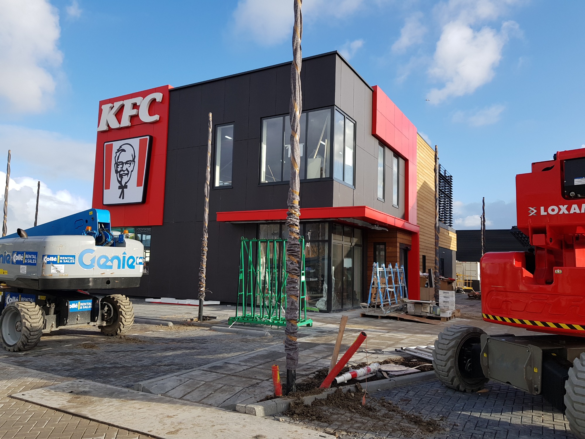 Nowoczesny budynek KFC w trakcie wykańczania elewacji, widoczne podnośniki koszowe i materiały budowlane na zewnątrz.