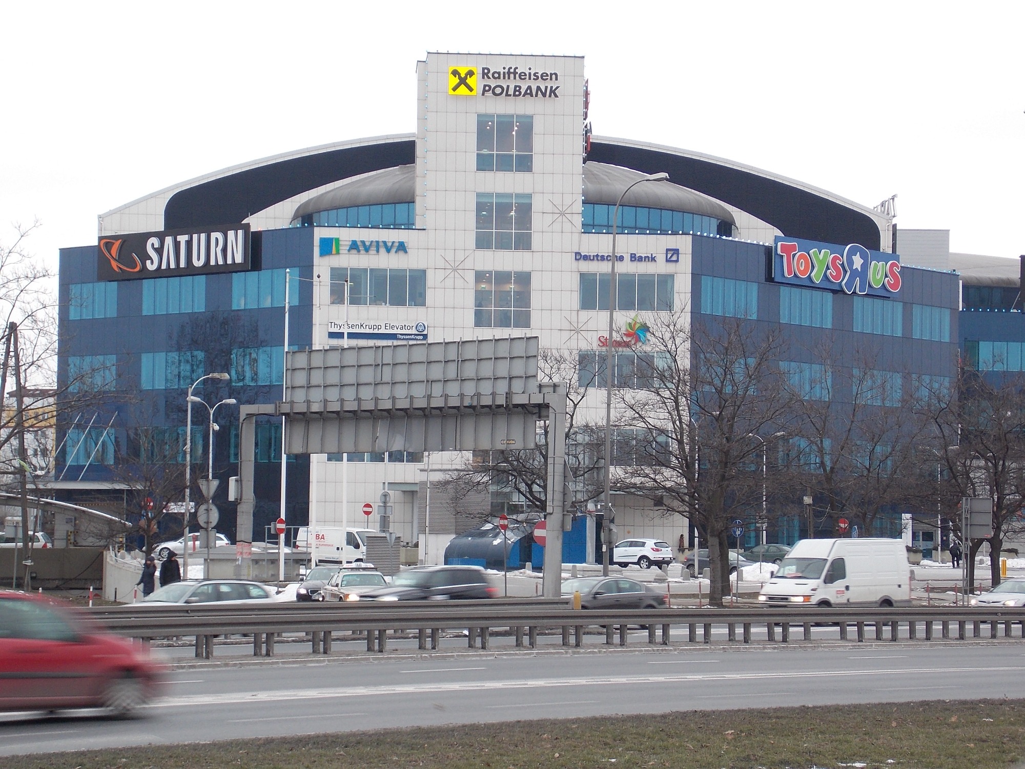 Wielopiętrowy budynek biurowy z niebieską szklaną fasadą i widocznymi szyldami firm Raiffeisen Polbank, Saturn, Aviva, Deutsche Bank i Toys'R'Us, sfotografowany z perspektywy ulicy...