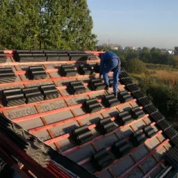 Dekarz w niebieskim kombinezonie układa czarną dachówkę karpiówkę na łatach pokrytych membraną dachową, widok z góry na częściowo pokryty dach i oddalony krajobraz.