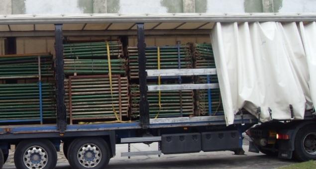 Ciężarówka z naczepą wypełniona metalowymi stemplami budowlanymi w kolorze zielonym i brązowym, zabezpieczonymi pasami transportowymi.