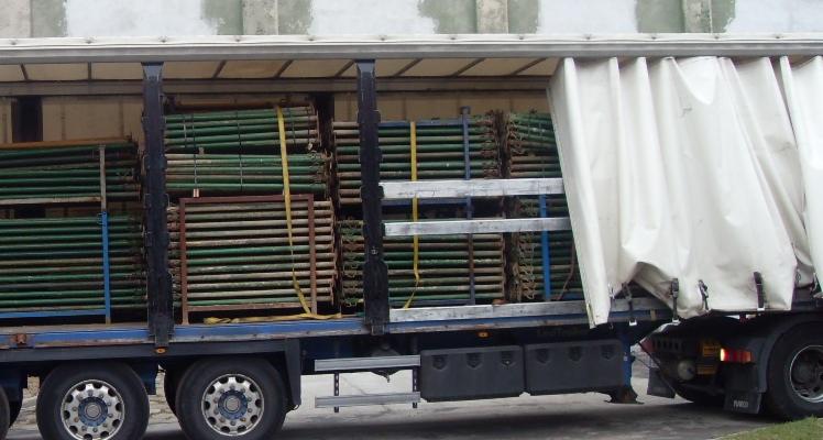 Ciężarówka z odsłoniętą plandeką, załadowana metalowymi stemplami budowlanymi, zabezpieczonymi żółtymi pasami.