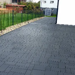 Nowoczesny podjazd z ciemnoszarej kostki brukowej ułożonej we wzór, obrzeża z jasnych kamieni, w tle siatka ogrodzeniowa i budynki.