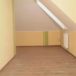 Poddasze z oknem dachowym, świeżo wykończone, z panelami podłogowymi o wzorze drewna i żółtymi ścianami z zielonym paskiem. Widoczny biały grzejnik.