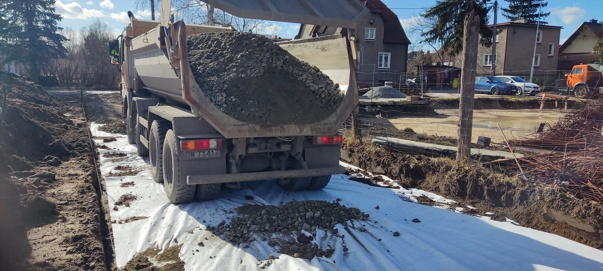 Ciężarówka wyładowuje gruz na geowłókninę w wykopie, widoczne opony, błoto i budynki w tle.